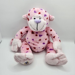 May include: A pink plush monkey toy, decorated with red and purple heart shapes. The monkey has a white face, hands, and feet, and a purple nose. It is sitting on a white surface.