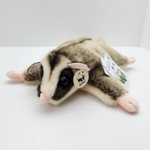 May include: A plush sugar glider toy with soft grey and white fur, dark brown facial markings, and pink paws. The toy features a round beige tag with a black panda logo and the text WWF, along with a white rectangular paper tag attached to its back.