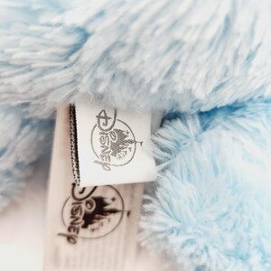 Peut inclure: Gros plan sur un jouet en peluche bleu clair avec une &eacute;tiquette Disney. L'&eacute;tiquette pr&eacute;sente le logo et le texte Disney. La peluche a une texture douce et moelleuse. L'&eacute;tiquette est blanche avec du texte noir et une bordure noire.