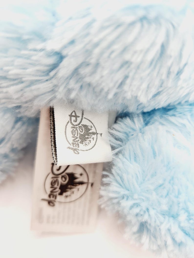 Peut inclure: Gros plan sur un jouet en peluche bleu clair avec une &eacute;tiquette Disney. L'&eacute;tiquette pr&eacute;sente le logo et le texte Disney. La peluche a une texture douce et moelleuse. L'&eacute;tiquette est blanche avec du texte noir et une bordure noire.