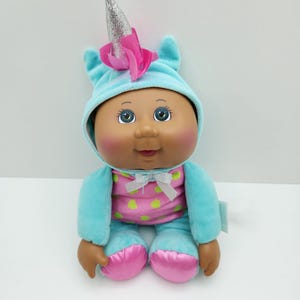 May include: A Cabbage Patch Kids doll wearing a blue unicorn-themed outfit. The doll has brown skin, blue eyes, and a pink and green polka-dotted shirt. The unicorn horn is silver glitter with a pink flower.