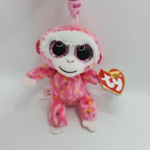May include: Pink and white monkey plush keychain with large sparkly eyes. The monkey is wearing a pink and white patterned outfit. The keychain has a pink plastic clip.