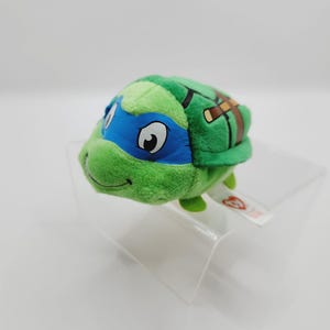 May include: Green and blue Teenage Mutant Ninja Turtles plush toy with a brown bandana and a red beanie. The toy is sitting on a clear acrylic stand.