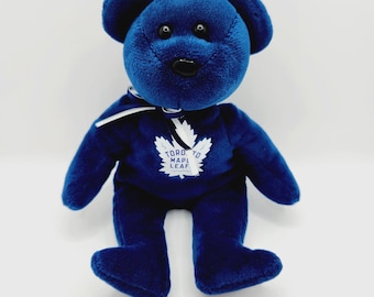 May include: A dark blue plush teddy bear with black plastic eyes and a black nose. The bear features a white ribbon tied around its neck and a white Toronto Maple Leaf logo printed on its chest with the text TORONTO MAPLE LEAF.
