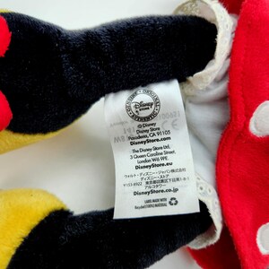 May include: A plush toy featuring black limbs and a red garment with white polka dots and white lace trim. A white fabric tag is attached, displaying the Disney Store logo, addresses in Pasadena, London, and Tokyo, and the text Label made with recycled (100%) material.