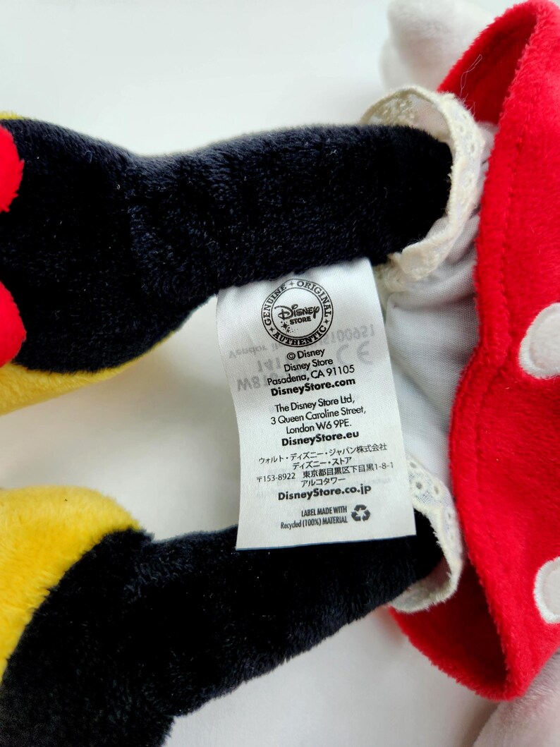 May include: A plush toy featuring black limbs and a red garment with white polka dots and white lace trim. A white fabric tag is attached, displaying the Disney Store logo, addresses in Pasadena, London, and Tokyo, and the text Label made with recycled (100%) material.