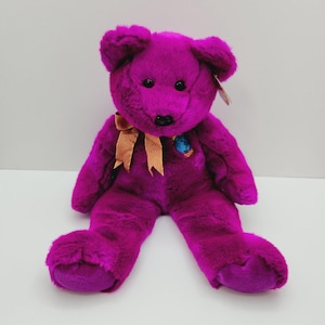 Peut inclure: Un ours en peluche violet avec un ruban brun autour du cou. L'ours a un petit patch de globe sur sa poitrine avec l'année 2000 imprimée dessus.