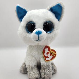 May include: A plush white cat toy with large, glittery blue eyes and blue accents on the ears and nose. The stuffed animal has gray paws and a red heart-shaped tag with the brand name.