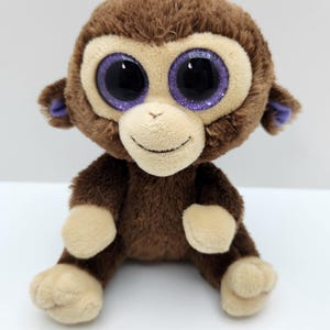 May include: A plush monkey toy with brown fur, a cream-coloured face, and large, glittery purple eyes. The monkey has small ears with purple inner accents, a stitched smile, and cream-coloured hands and feet. It is seated in an upright position.
