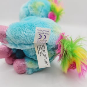 Op de afbeelding: Een klein, blauw en groen pluche eenhoorn speelgoed met een roze manen en staart. Het speelgoed heeft een label met de tekst "New Material Only Reg. No. 02T-00454211 Content: Polyester fiber and Plastic beads Handmade in China 0112 Reg. No. PA-15626(CN) CE & Plastic Pellets"