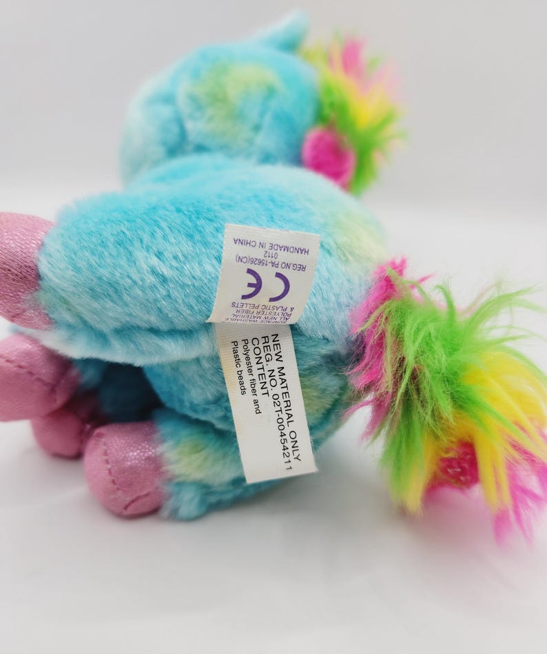 Op de afbeelding: Een klein, blauw en groen pluche eenhoorn speelgoed met een roze manen en staart. Het speelgoed heeft een label met de tekst "New Material Only Reg. No. 02T-00454211 Content: Polyester fiber and Plastic beads Handmade in China 0112 Reg. No. PA-15626(CN) CE & Plastic Pellets"
