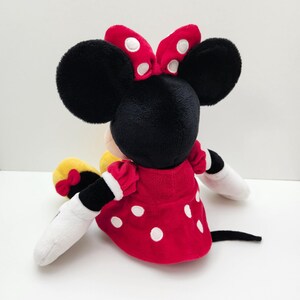 May include: A plush toy of a cartoon mouse character with large black ears, wearing a red dress with white polka dots and a matching red bow with white dots between the ears. The character has white gloves, yellow shoes with small red bows, and a black tail.