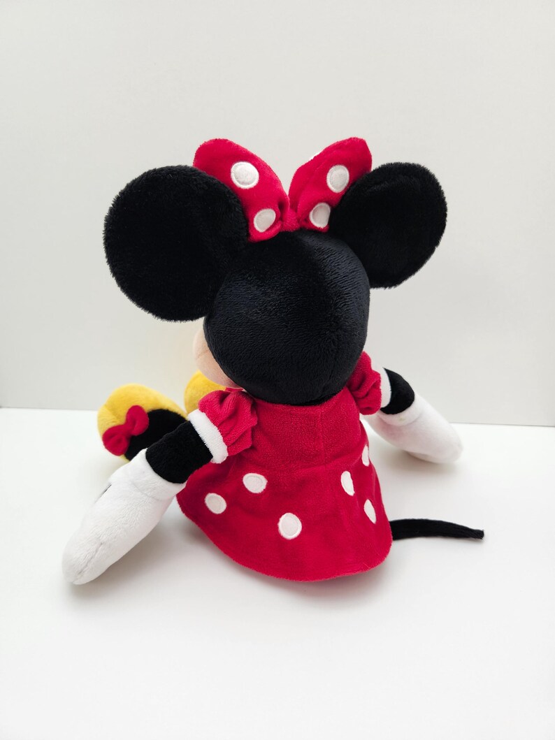 May include: A plush toy of a cartoon mouse character with large black ears, wearing a red dress with white polka dots and a matching red bow with white dots between the ears. The character has white gloves, yellow shoes with small red bows, and a black tail.