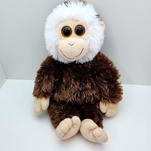 May include: A plush monkey toy with a brown body and a white furry head. The toy has large, round eyes and a smiling face. The monkey is sitting on a white surface.