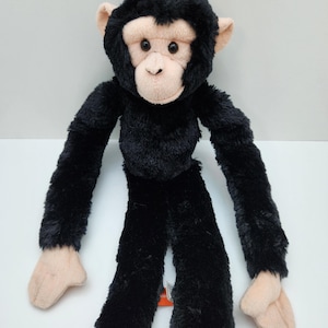 May include: A plush chimpanzee toy featuring soft black fur covering its body, head, and long arms. The face, ears, and hands are made of a light beige fabric. The chimpanzee has dark, round eyes and a neutral expression, with long limbs.