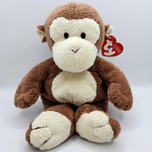 May include: A plush monkey toy with brown fur and cream-colored accents on its face, chest, and feet. The stuffed animal has a red heart-shaped tag attached to its ear.