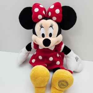 May include: A plush Minnie Mouse toy featuring a red dress with white polka dots, a matching red bow with white polka dots, black ears, white gloves, and yellow shoes. The bottom of the left yellow shoe features a circular gold Disney patch.