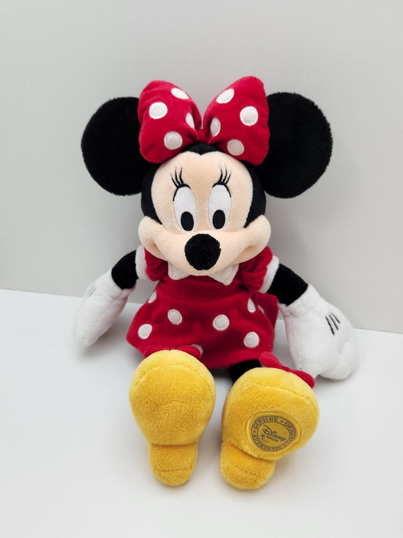 May include: A plush Minnie Mouse toy featuring a red dress with white polka dots, a matching red bow with white polka dots, black ears, white gloves, and yellow shoes. The bottom of the left yellow shoe features a circular gold Disney patch.