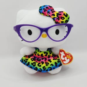 May include: A white plush toy cat wearing purple glasses and a colorful bow and dress with a leopard print design. The toy has black eyes and a yellow nose. A red heart-shaped tag is attached.