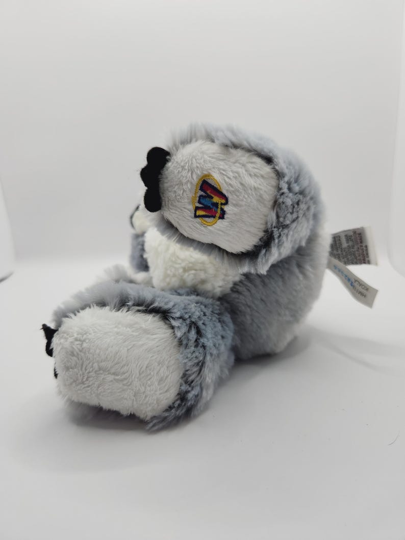 May include: A gray and white plush toy with a black nose and a Webkinz tag. The toy is sitting on a white surface.