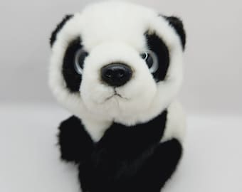 May include: A plush panda bear with a black nose, black ears, and black patches around the eyes and on the limbs. The panda's fur is white and fluffy, and it has large, light blue eyes. The panda is sitting upright.