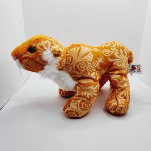 May include: A plush tiger toy in orange with a white floral pattern and white fur around the neck. The toy has a pink nose and black eyes. A small tag with the word "GANZ" is attached to the toy.