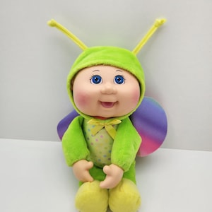 May include: A Cabbage Patch Kids doll dressed in a lime green insect costume with antennae, a polka-dot chest panel, a yellow ribbon bow, and iridescent purple and blue wings. The doll has blue eyes, rosy cheeks, and a smiling expression.