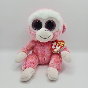 May include: Pink and white plush monkey with large sparkly eyes. The monkey is sitting with its legs crossed and has a pink and yellow floral pattern on its body. The monkey has a Ty Beanie Boo tag attached to its arm.