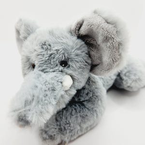 Może przedstawiać: Pluszowa szara zabawka w kształcie słonia z dużymi uszami, trąbą i małymi czarnymi oczami. Zabawka leży na białej powierzchni. Sierść słonia jest jasnoszara.