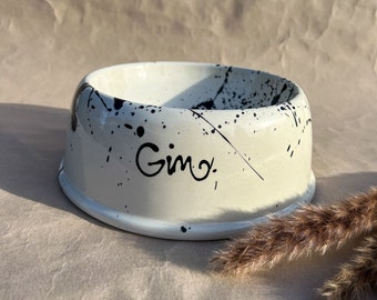 Puede incluir: Tazón de cerámica blanco para mascotas con salpicaduras de pintura negra y el nombre "Gimo" escrito en cursiva negra.