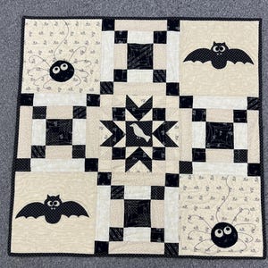 May include: A black and white quilt with a Halloween theme. The quilt features bats, spiders, and a bird in a star-shaped design.