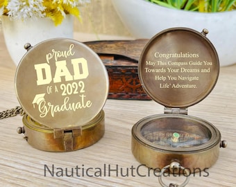 May include: A brass compass with a gold inscription that reads "Congratulations May This Compass Guide You Towards Your Dreams and Help You Navigate Life' Adventure." The compass is open and the needle is pointing north. The compass is attached to a gold chain. The compass is sitting on a wooden surface. The inscription on the other side of the compass reads "Proud Dad of a 2022 Graduate".