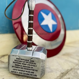 Può includere: Una replica in argento del martello Mjolnir con un manico in pelle marrone e un'iscrizione che recita "FOR INSTANCE, GOD TEACHES US TP LOVE BY PUTTING SOME UNLOVELY PEOPLE AROUND US. IT TAKES NO CHARACTER TO LOVE PEOPLE WHO ARE LOVELY AND LOVING TO YOU". Il martello è davanti a uno scudo di Capitan America rosso, bianco e blu.