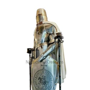 Puede incluir: Una armadura completa de plata con una capa blanca, un casco con una cruz y un escudo con una cruz y un caballero a caballo. El escudo tiene el texto "XPSTI MILITVM" alrededor de la imagen.