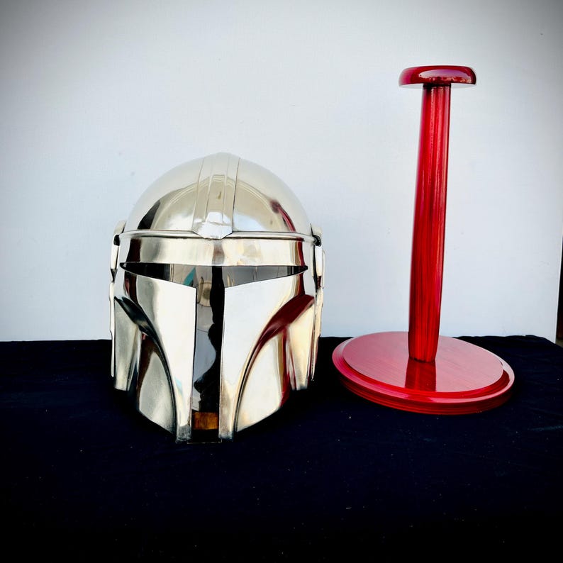 May include: A silver Mandalorian helmet with a red wooden display stand. The helmet has a visor and a textured surface.