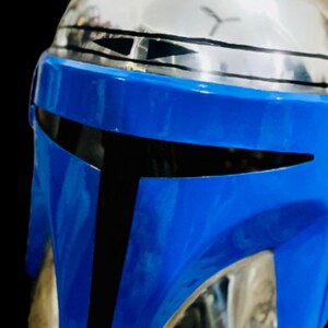May include: A blue and black Mandalorian helmet with a silver visor. The helmet has a black stripe across the top and a black stripe across the bottom.