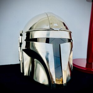 May include: A silver Mandalorian helmet with a black visor. The helmet is made of metal and has a smooth, polished finish.