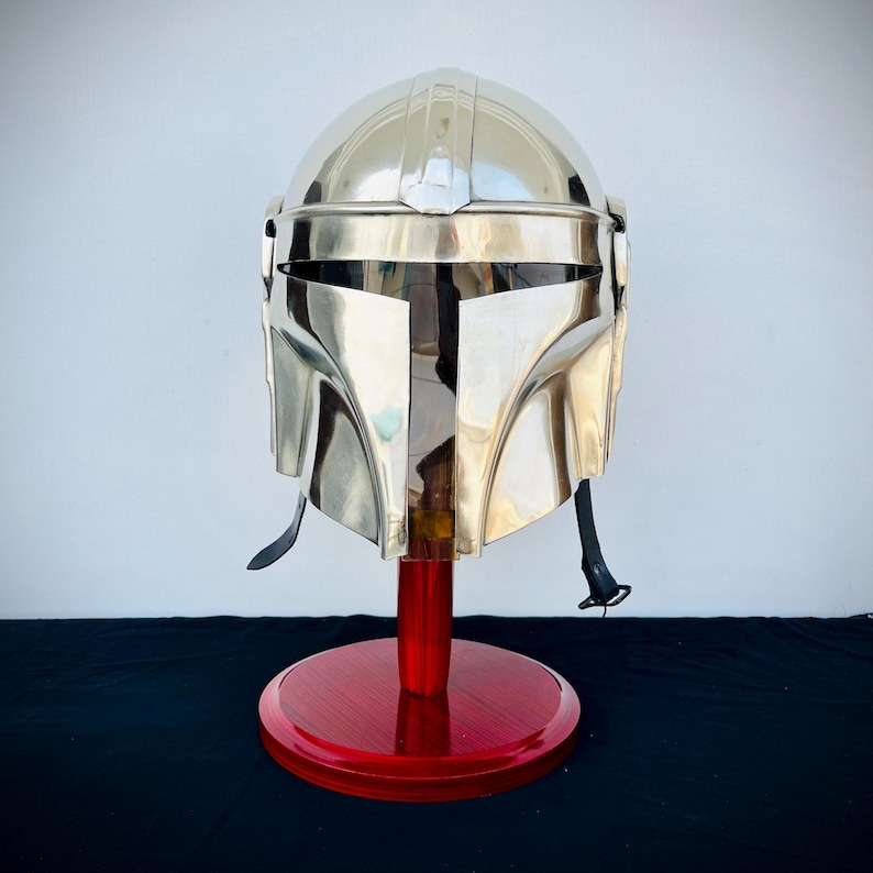 May include: A silver Mandalorian helmet with a red wooden display stand. The helmet has a visor and a T-shaped design on the front.