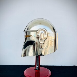 May include: A silver Mandalorian helmet with a red wooden stand. The helmet has a yellow visor and intricate details.