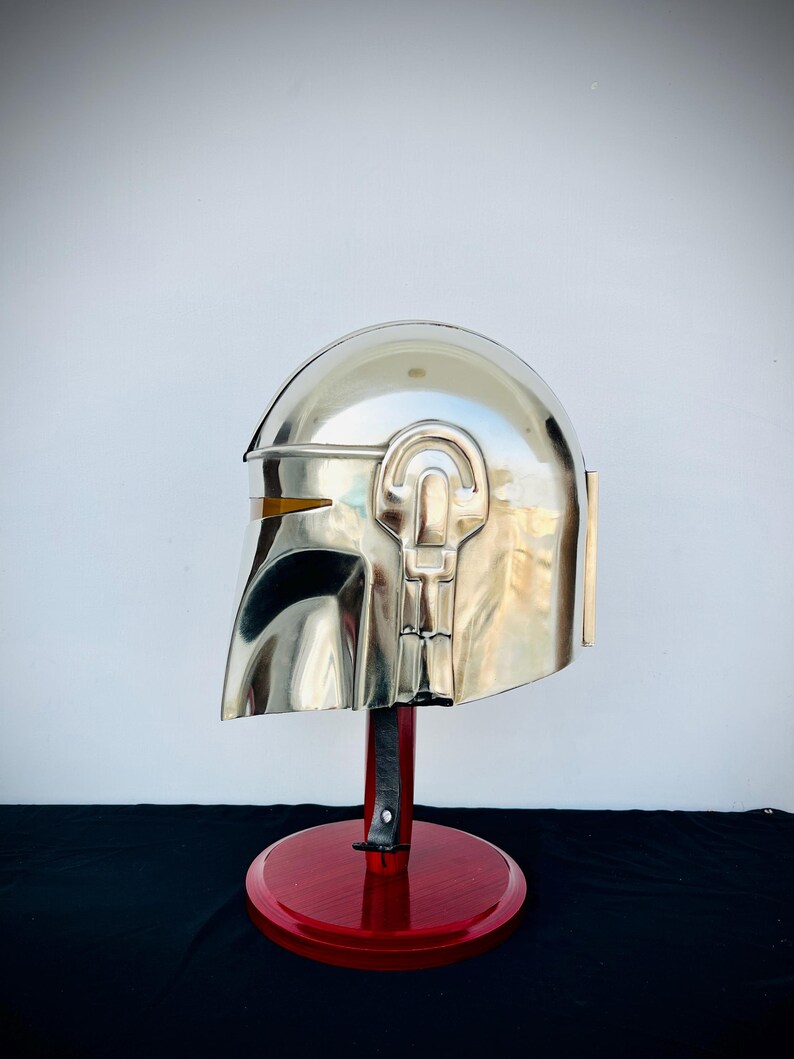 May include: A silver Mandalorian helmet with a red wooden stand. The helmet has a yellow visor and intricate details.