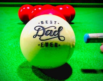 May include: A white billiard ball with the text "Best Dad Ever" printed in black on the ball. The ball is on a green felt pool table with red billiard balls in the background.