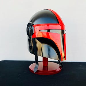 May include: A black and red helmet with a visor, displayed on a red stand. The helmet has a textured surface and a sleek design.