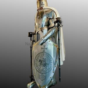 Puede incluir: Una armadura completa de plata con una capa blanca, un casco con una cruz y un escudo con una cruz y un caballero a caballo. El escudo tiene el texto "XPSTI MILITVM SVB SIBI" alrededor del caballero.