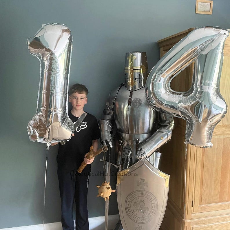 Puede incluir: Un ni&ntilde;o con una camisa negra sostiene un globo plateado n&uacute;mero uno. Un globo plateado n&uacute;mero cuatro est&aacute; en el fondo. Un caballero con armadura se encuentra detr&aacute;s del ni&ntilde;o, sosteniendo un escudo con un escudo de armas.