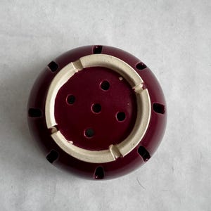Op de afbeelding: Een bordeauxrode keramische wierookbrander met een cr&egrave;mekleurige binnenrand. De brander heeft verschillende kleine gaten aan de zijkanten en een reeks gaten in het midden. De wierookbrander is ontworpen om wierookstokjes in te plaatsen.
