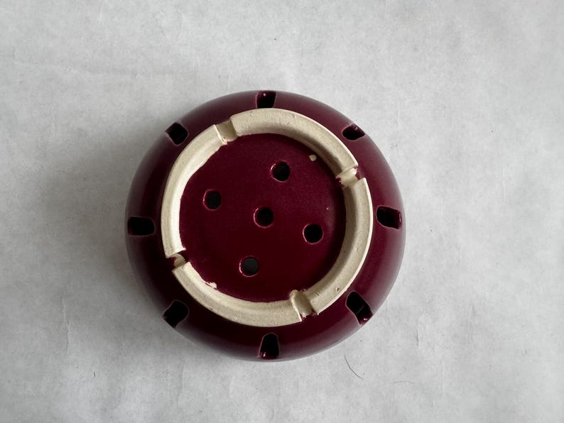 Op de afbeelding: Een bordeauxrode keramische wierookbrander met een cr&egrave;mekleurige binnenrand. De brander heeft verschillende kleine gaten aan de zijkanten en een reeks gaten in het midden. De wierookbrander is ontworpen om wierookstokjes in te plaatsen.