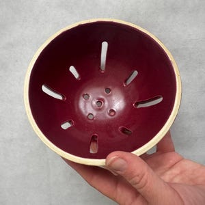May include: A ceramic berry bowl with a deep burgundy interior and a cream-colored rim. The bowl has several elongated holes around the sides and smaller holes in the center. This bowl is designed for washing and serving berries.