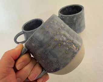 Op de afbeelding: Drie keramische mokken met een gespikkeld blauw en wit glazuur. De mokken hebben een lichtgrijze rand en een gespikkeld gouden glazuur.
