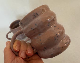 Op de afbeelding: Twee handgemaakte keramische mokken met een gespikkeld, lichtbruin glazuur. De mokken hebben een uniek, golvend ontwerp en een afgeronde handgreep.