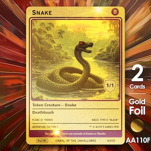 以下が含まれることがあります： 沼地環境にいるヘビが描かれた金色のトレーディングカード。「SNAKE」と書かれ、「Token Creature - Snake」と「Deathtouch」の文字が入っています。カードには1/1のシンボルと「Adventure Edition II」の文字も入っています。